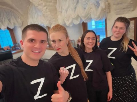 Młody mężczyzna oraz trzy młode kobiety pozują do zdjęcia w czarnych koszulkach z białą literą Z. Biała litera Z malowana jest na pojazdach wojskowych Federacji Rosyjskiej biorących udział w inwazji na Ukrainę.